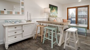 Una habitación luminosa y bien iluminada con un aparador blanco, una mesa de madera y dos taburetes. Un espejo grande y un cuadro adornan la pared.