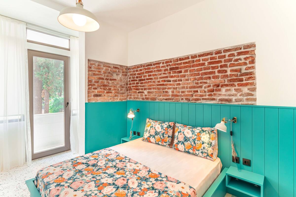 A bedroom with a brick wall, teal painted walls, a floral bedspread, and a sliding glass door to a balcony.