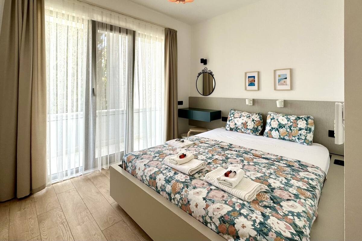 A bedroom with a large bed, a bedside table, a mirror, and a view of the outdoors through the window.