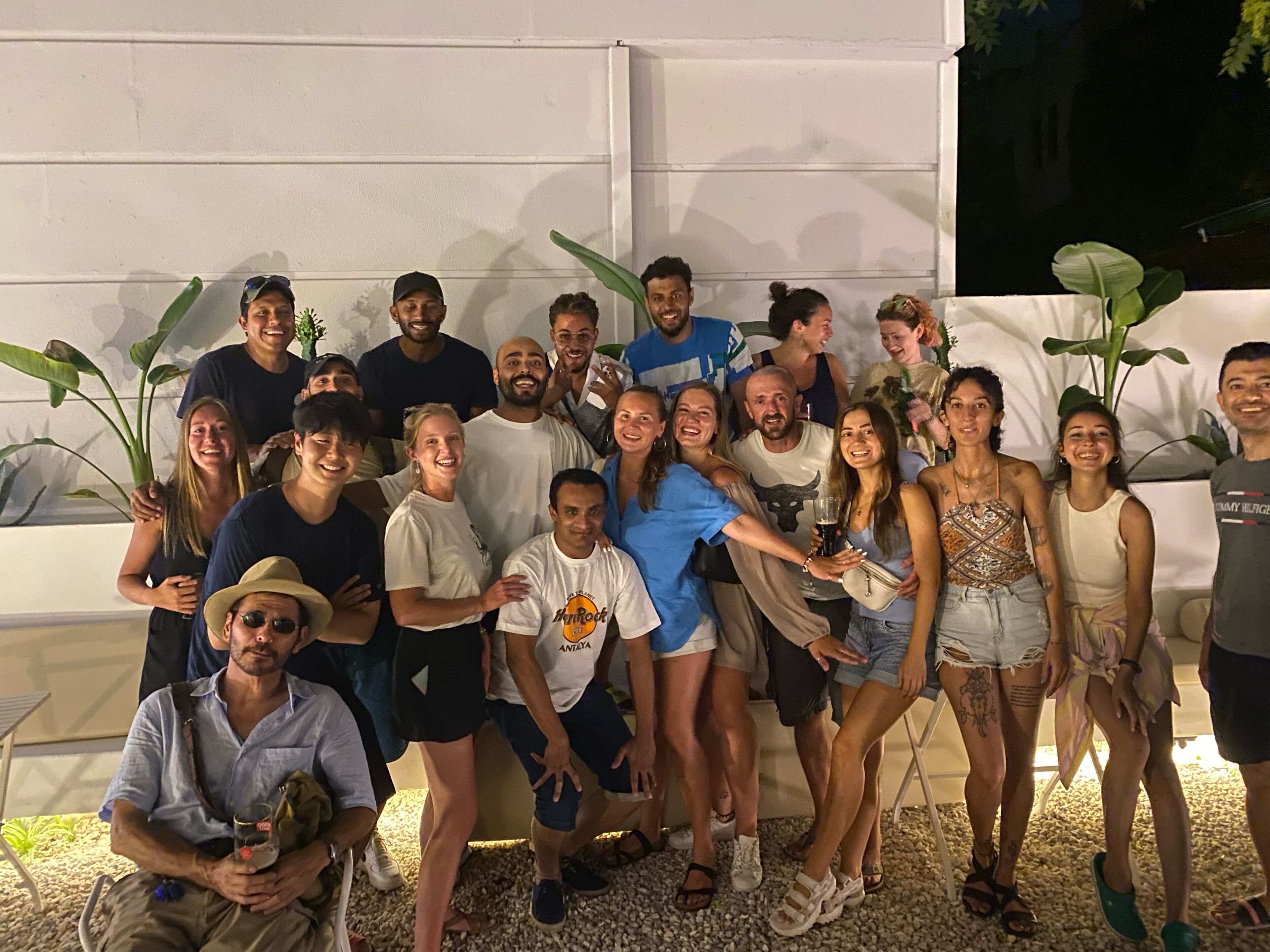 Un grupo de personas posando en un entorno exterior con un muro blanco y plantas verdes grandes al fondo.