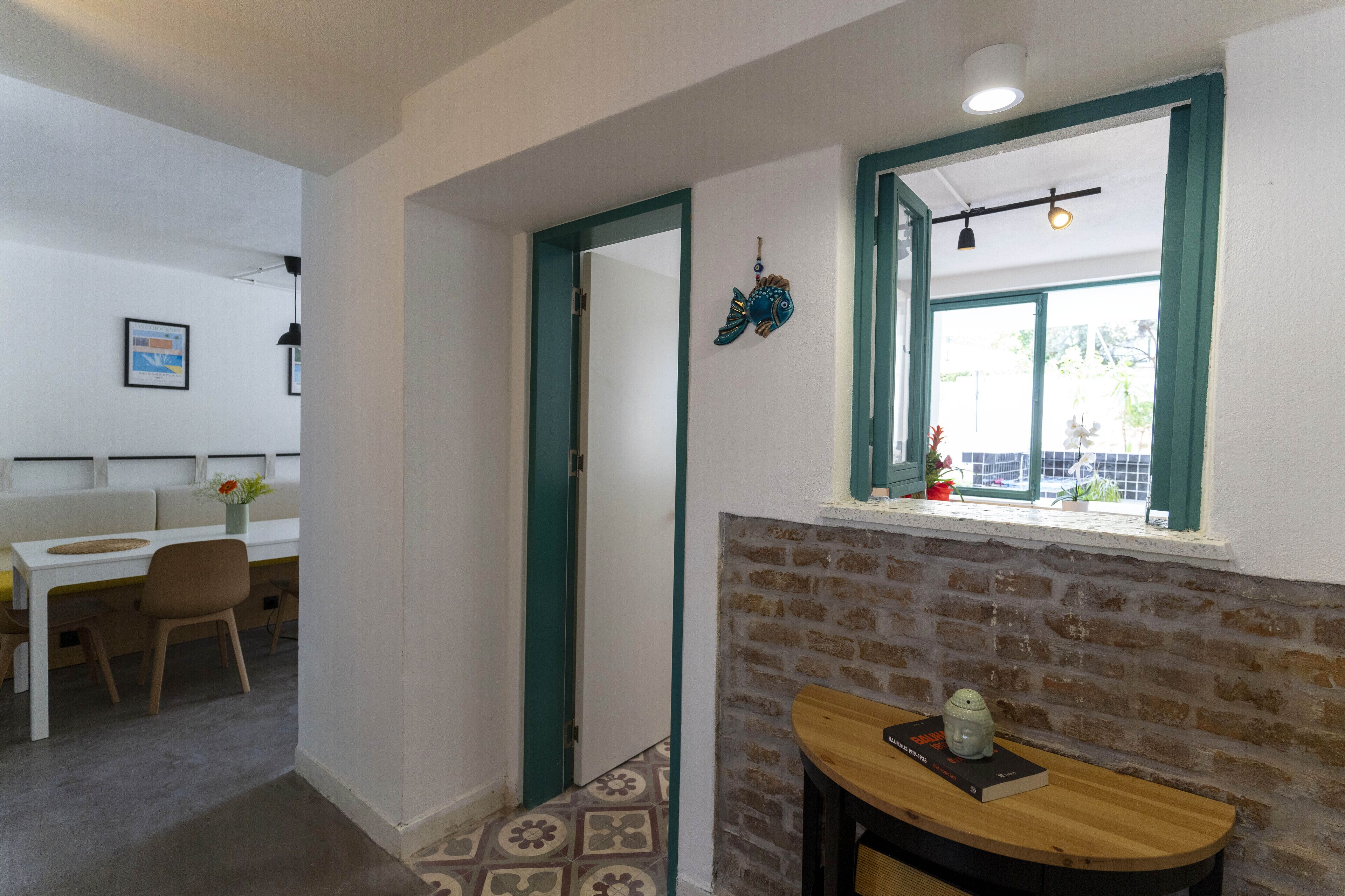 A room with a brick wall, a window with a view, a table with a book, and a door leading to another room.