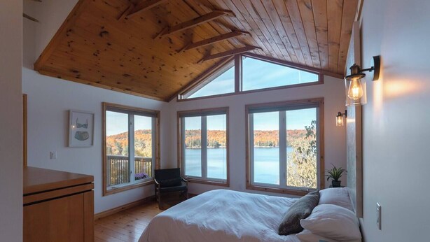 A bedroom with a large bed, wooden ceiling, and multiple windows offering a view of a lake and autumn foliage. - Rustic-Luxury Waterfront Cottage Getaway on Kennisis Lake Stunning Elevated View (Haliburton)