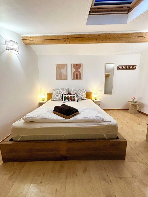 A modern bedroom with a wooden platform bed, white bedding, two abstract wall art pieces, a mirror, and a small bedside table with a lamp.