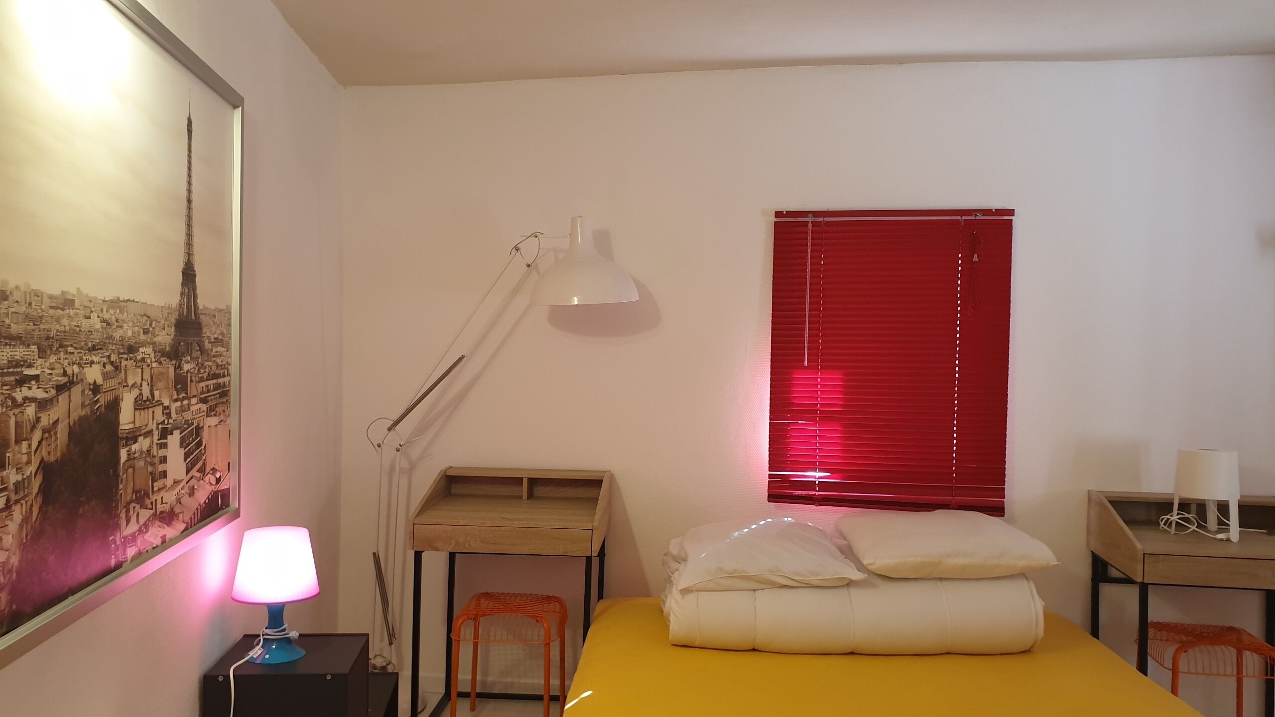 A hotel room with a bed, a desk, a chair, a lamp, a window with blinds, and a painting on the wall.