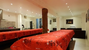 Une chambre d’hôtel dotée d’une longue table à manger recouverte d’une nappe rouge, d’un coin cuisine avec une cuisinière, un réfrigérateur et un évier, et d’un grand pilier au centre.
