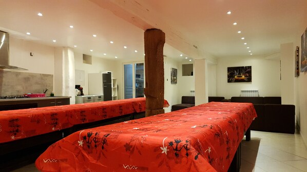 Une chambre d’hôtel dotée d’une longue table à manger recouverte d’une nappe rouge, d’un coin cuisine avec une cuisinière, un réfrigérateur et un évier, et d’un grand pilier au centre.