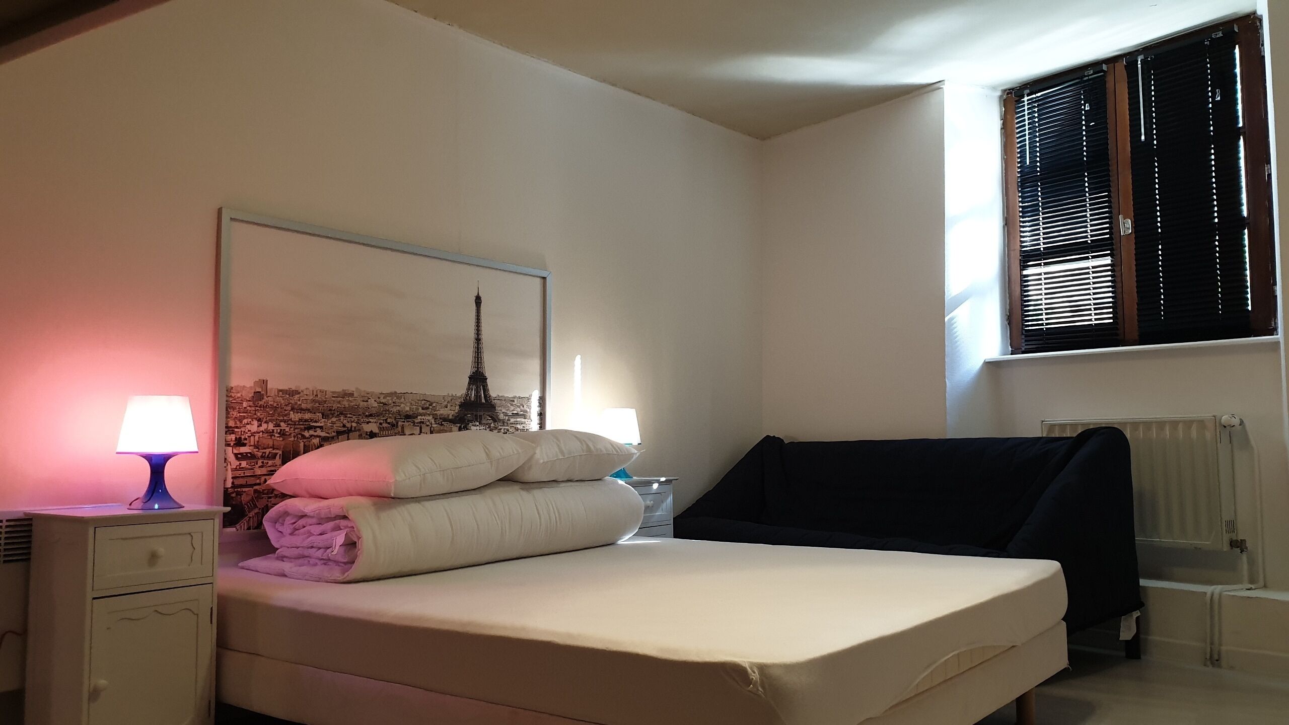 A bedroom with a bed, a nightstand, a lamp, a window with blinds, and a wall-mounted radiator.