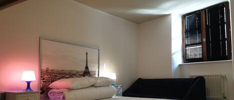 A bedroom with a bed, a nightstand, a lamp, a window with blinds, and a wall-mounted radiator.