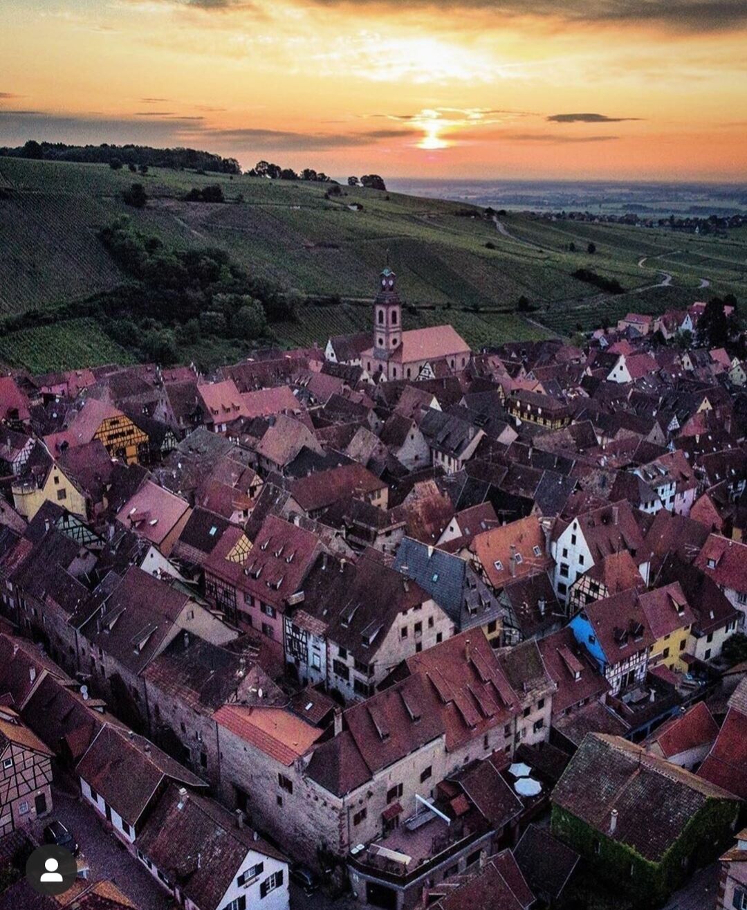 Un pueblo histórico con edificios apretados, una iglesia destacada con un campanario y un atardecer al fondo.