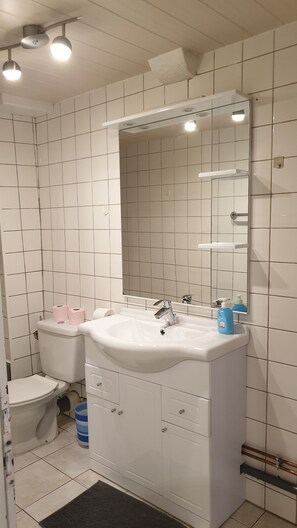 Une salle de bain avec des carreaux blancs, un lavabo blanc, un miroir et des toilettes.