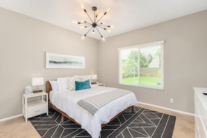 A bedroom with a bed, bedside tables, a nightstand, a ceiling light fixture, a window with blinds, and a painting on the wall.