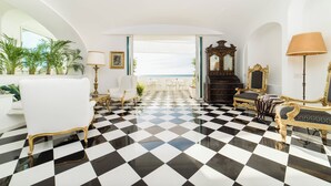 Een kamer met een zwart-wit schakerend vloerkleed, wit en goudkleurig meubilair en uitzicht op zee door een groot raam.