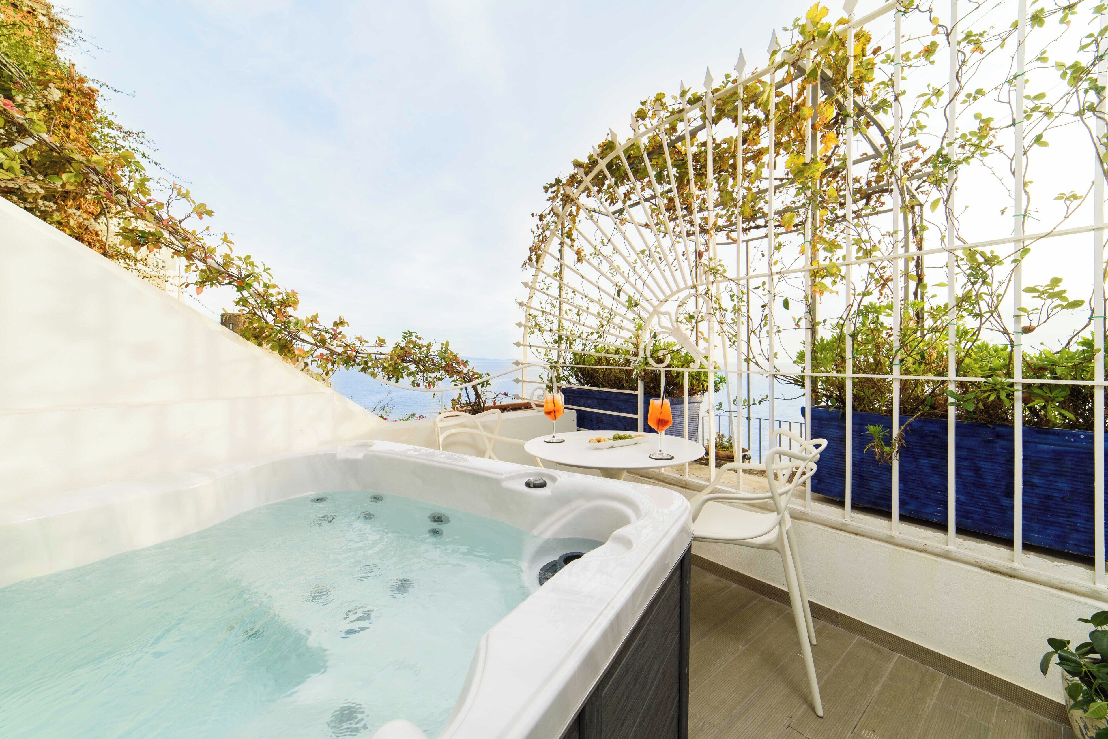 Uma banheira de hidromassagem com vista para o mar, uma área de estar exterior em branco e uma pérgula coberta por plantas trepadeiras.