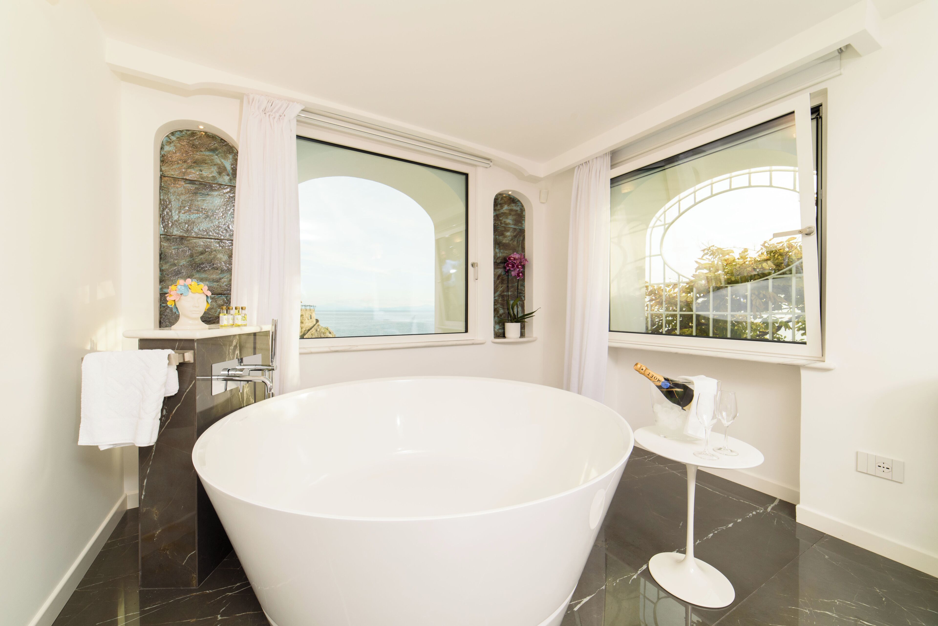 Un baño moderno con una bañera independiente grande, una mesita con una botella y un vaso, y ventanas amplias con vista al mar.
