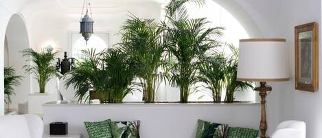 Een moderne woonkamer met een witte bank, groene kussens met een patroon, een vloerlamp en een groot raam met planten.