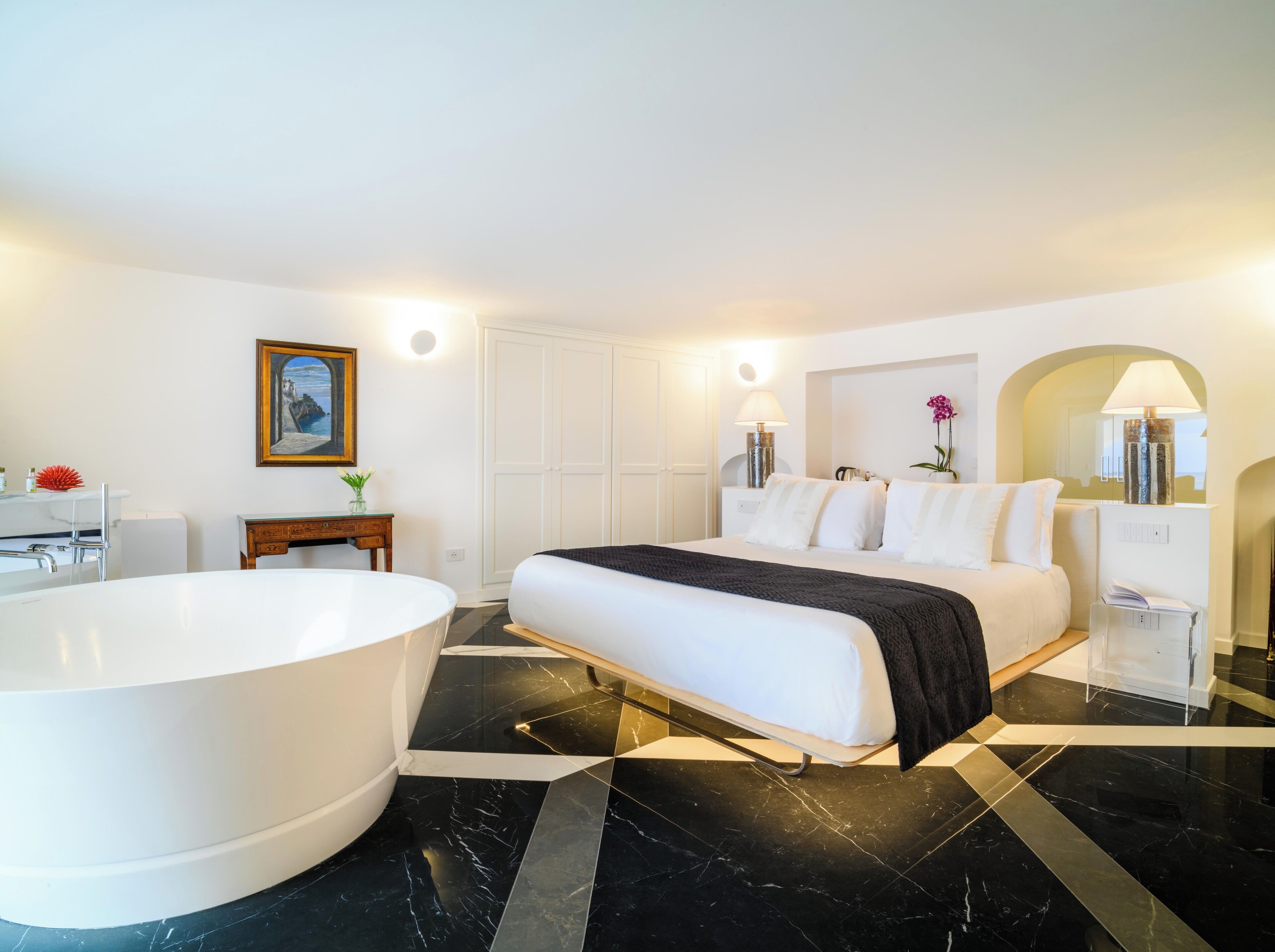 A modern hotel room with a large bed, a bathtub, a vanity with a mirror, and a painting on the wall.