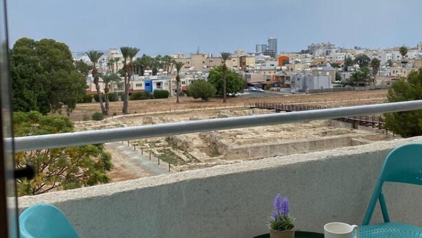 A balcony with two turquoise chairs, a small table with a white mug, a glass of water, and a potted plant. The view includes a cityscape with buildings and palm trees. - Larnaca Comfort Rooms (Larnaca)