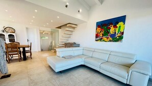 A modern living room with a white leather sectional sofa, a dining area with wooden chairs, a staircase, and a colorful abstract painting on the wall. - Villa a schiera Punta Aldia (Puntaldia)