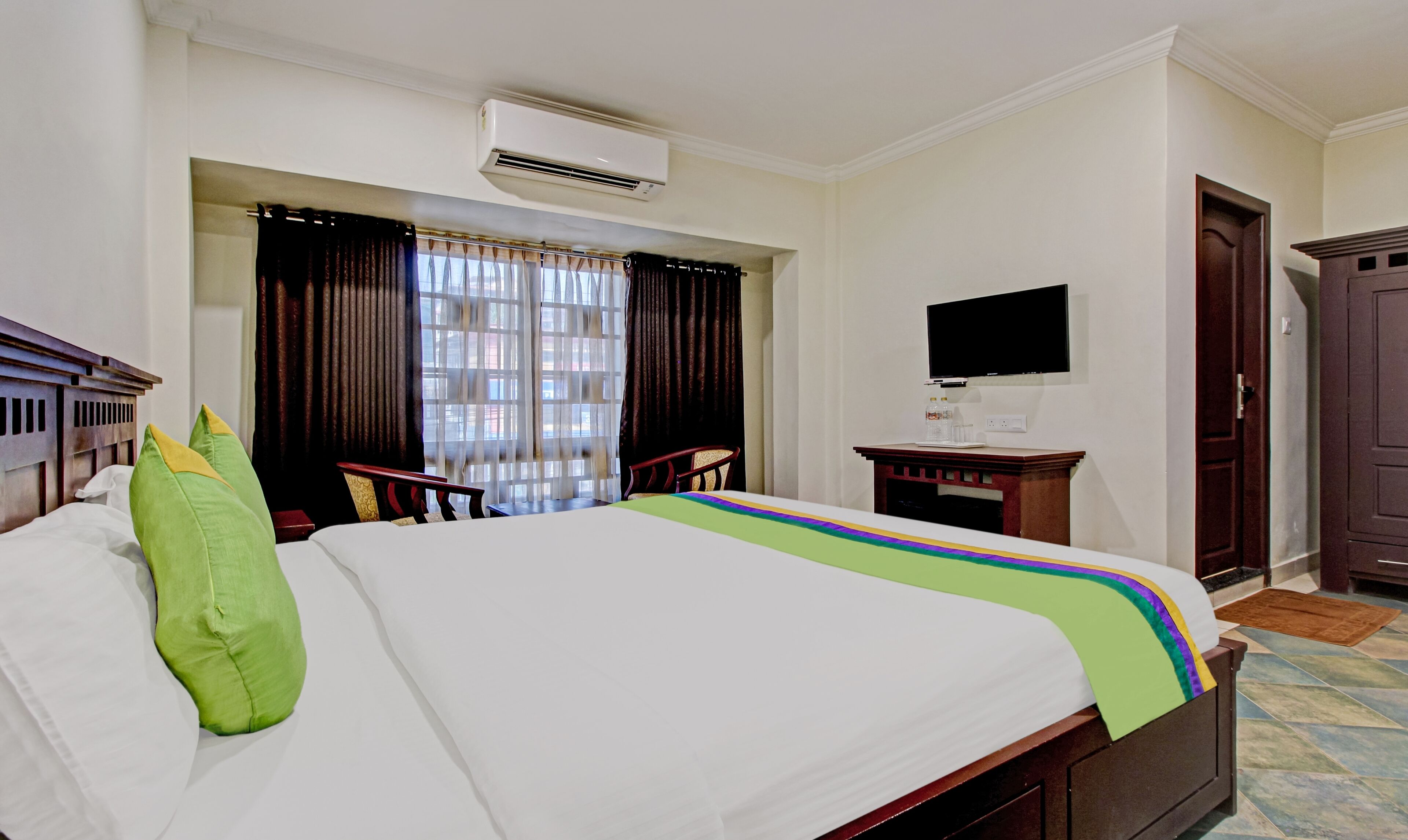 A hotel room with a bed, a TV, an air conditioning unit, a desk with a chair, and a door.