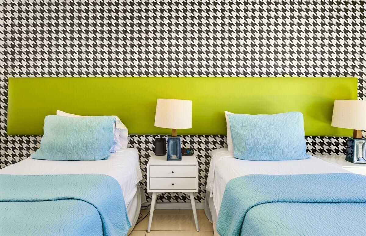Een hotelkamer met twee bedden, een groen hoofdeinde, nachtlampjes en een muur met een hondentandpatroon.