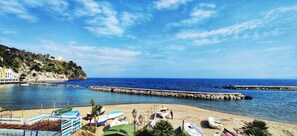 Una spiaggia costiera con un litorale roccioso, un molo che si protende verso il mare e diverse imbarcazioni sulla sabbia.