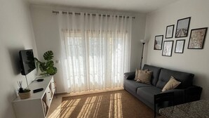 A modern living room with a sofa, a dining table with chairs, a TV, and a large window with sheer curtains. - Qta.da Flor - Fantastic Apartment With Pool (Albufeira)