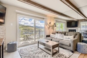 A spacious living room with a large sliding glass door leading to a balcony, a sofa with decorative pillows, a coffee table, and a flat-screen TV mounted on the wall. - Amazing Lake and Mountain Views! (Dillon)
