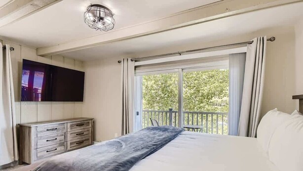 Ein Schlafzimmer mit Bett, einem an der Wand befestigten Fernseher, einer Holzkommode und einem Balkon mit Vorhängen.