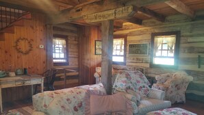 Interior rústico de una cabaña con una mesa de comedor de madera, un sofá estampado con motivos florales y un cartel que anuncia antigüedades y alfombras de gancho.