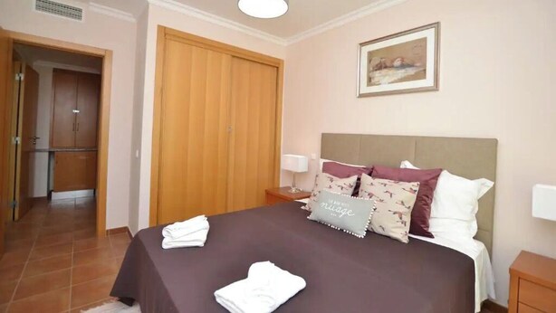 A hotel room with a bed, a nightstand, a lamp, a wooden wardrobe, and a painting on the wall. - Magnolia - Beach & Pool - Fantastic T2 (Albufeira)
