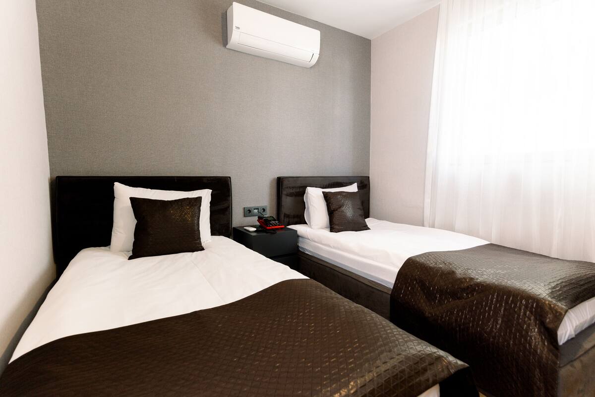A hotel room with two single beds, a nightstand with a lamp, and a wall-mounted air conditioner.