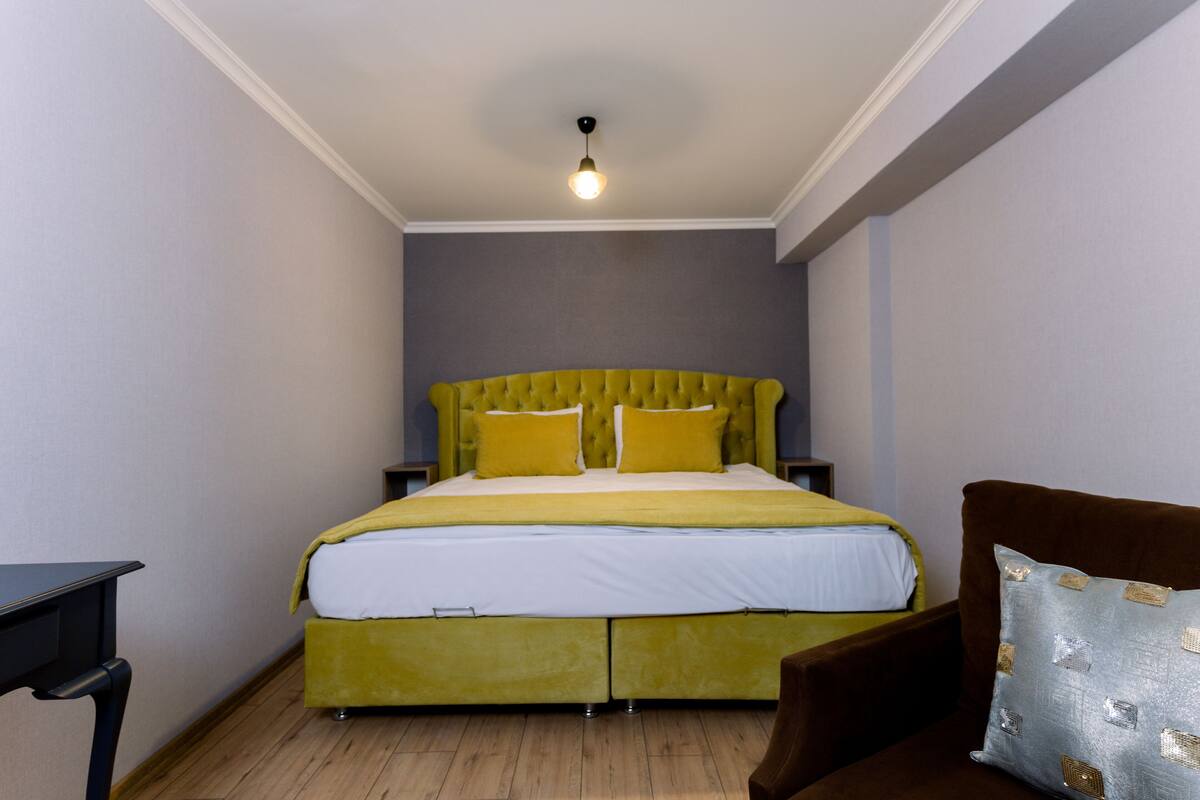 A bedroom with a green upholstered bed, two bedside tables, a wooden floor, and a single hanging light fixture.