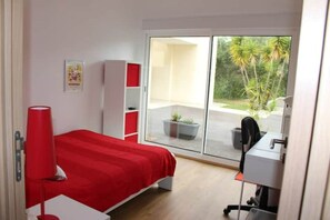 A bedroom with a bed, a desk, and a chair. There is a window with a view of palm trees and a balcony.
