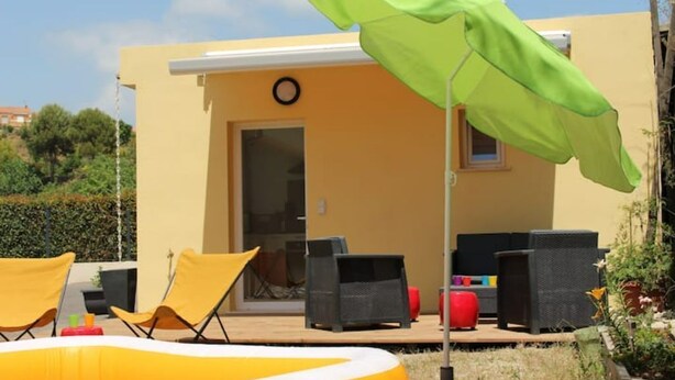 Un'area con posti a sedere all'aperto, con sedie gialle e un ombrellone verde, un edificio giallo con una porta e una finestra, e una piccola piscina gonfiabile.