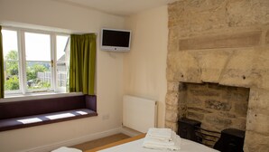 A bedroom with a bed, a window with curtains, a television, a stone fireplace, and a radiator. - The Star Inn (Stroud)