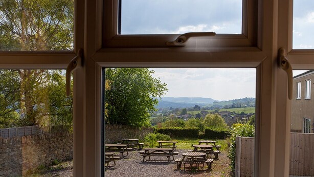 A view through a window of a patio area with multiple wooden tables and benches, surrounded by a stone wall and greenery. - The Star Inn (Stroud)
