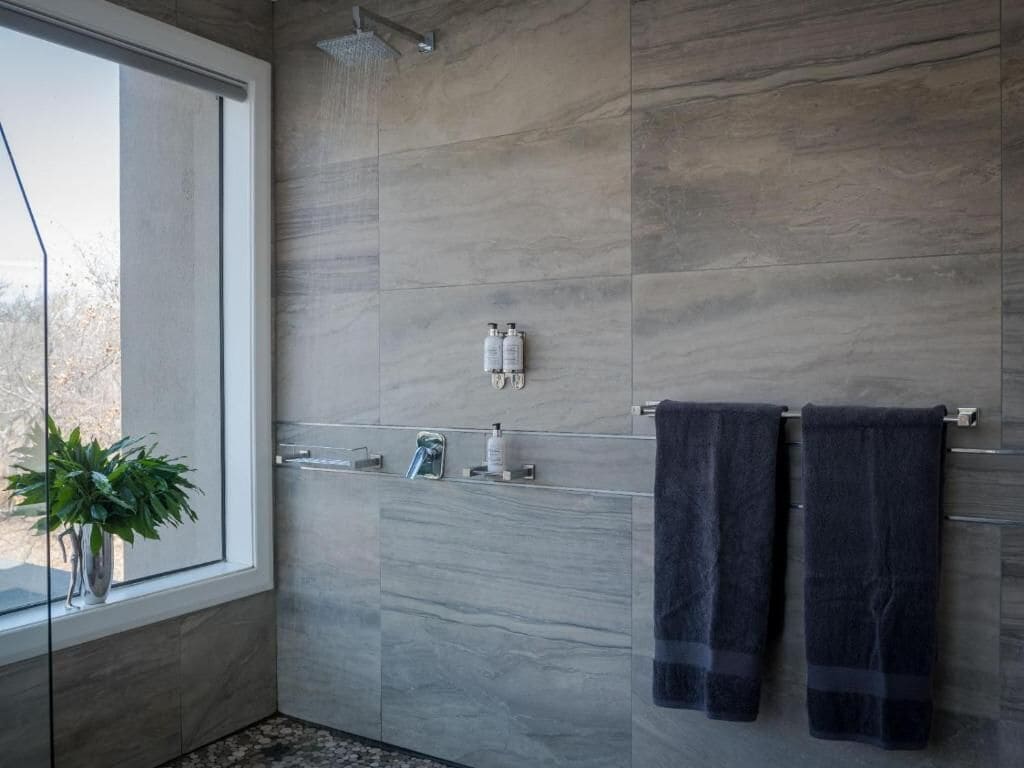 A modern bathroom with grey tiles, a glass shower enclosure, a towel rack with two towels, and a potted plant by the window.