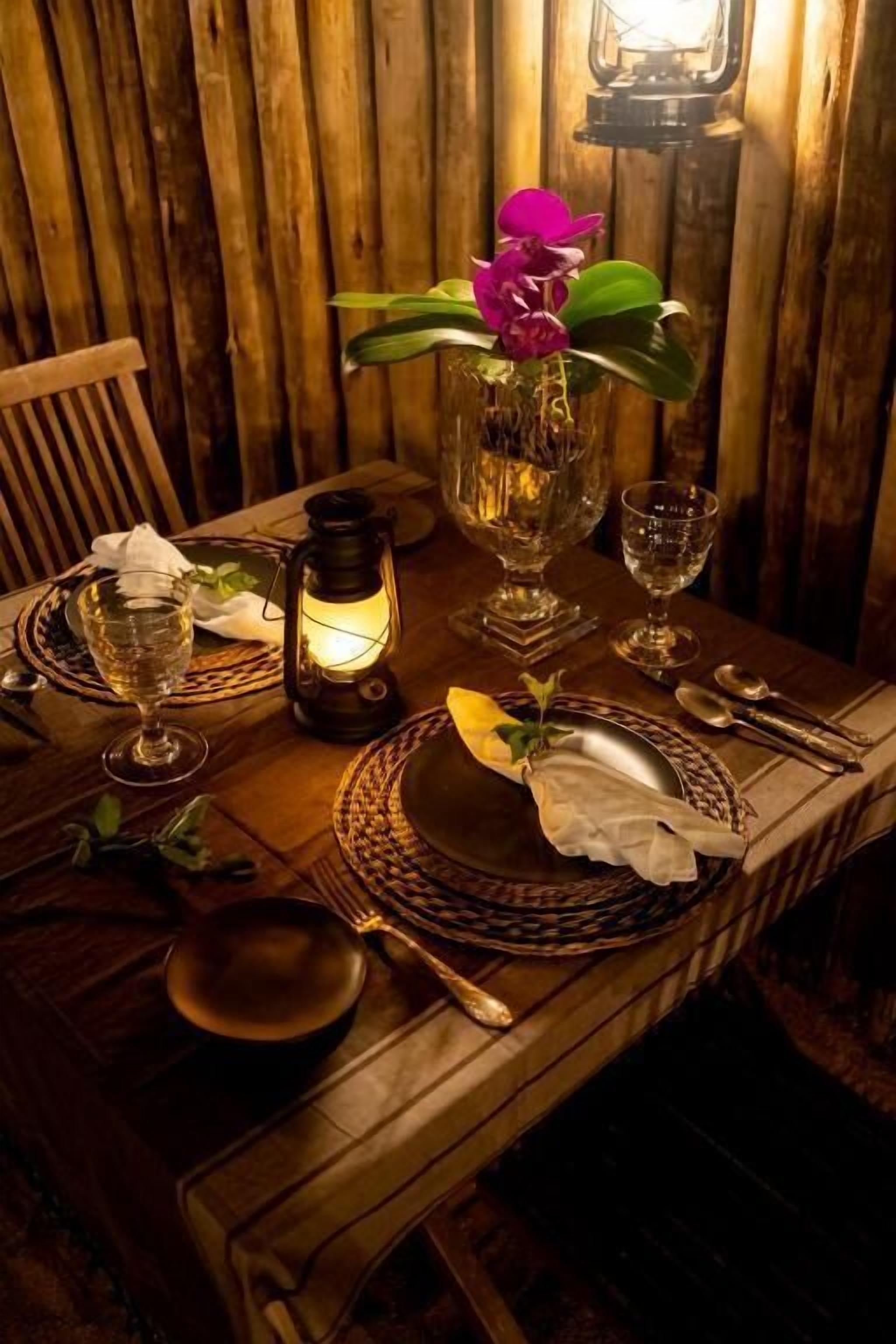 Mesa de jantar de madeira posta com uma vela, copos e um vaso com uma flor.