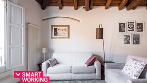 Una sala moderna con un sofá blanco, un sofá gris y un cojín decorativo estampado. Se observa un cuadro en la pared y una lámpara junto al sofá gris.