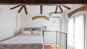 Un dormitorio con cama, pared de ladrillo curva, unidad de aire acondicionado, ventana con cortinas y suelo de madera.