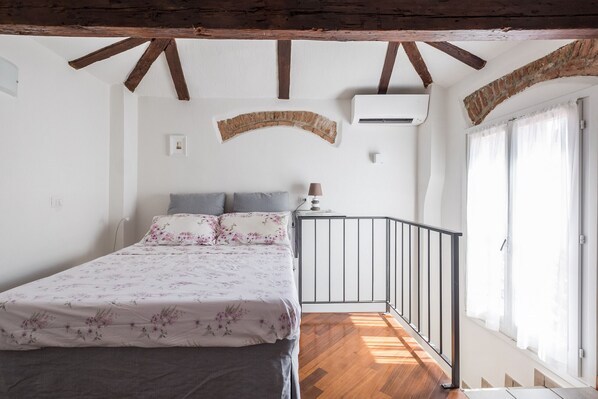 Ein Schlafzimmer mit einem Bett, einer gewölbten Ziegelwand, einer Klimaanlage, einem Fenster mit Vorhängen und einem Holzboden.