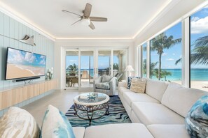 Un salón amplio con un televisor grande, asientos cómodos, un ventilador de techo y vista a la playa a través de amplias ventanas.