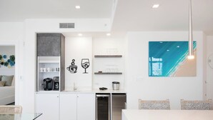 A modern kitchen with white cabinetry, a built-in oven, and a coffee machine. There is a dining area with a glass table and wicker chairs.