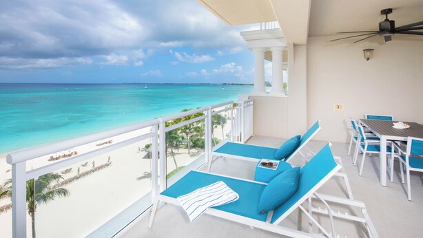 A balcony with a view of the ocean, white lounge chairs, a table with chairs, and a ceiling fan.