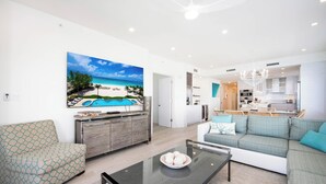 A modern living room with a large beach-themed painting, a sofa, a glass coffee table, and a kitchen area visible through an open door.