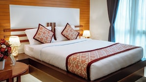 Quarto de hotel com cabeceira de madeira, uma cama grande com roupa de cama branca e almofadas estampadas, mesa de cabeceira com abajur e vista para o exterior através de uma janela com cortinas.