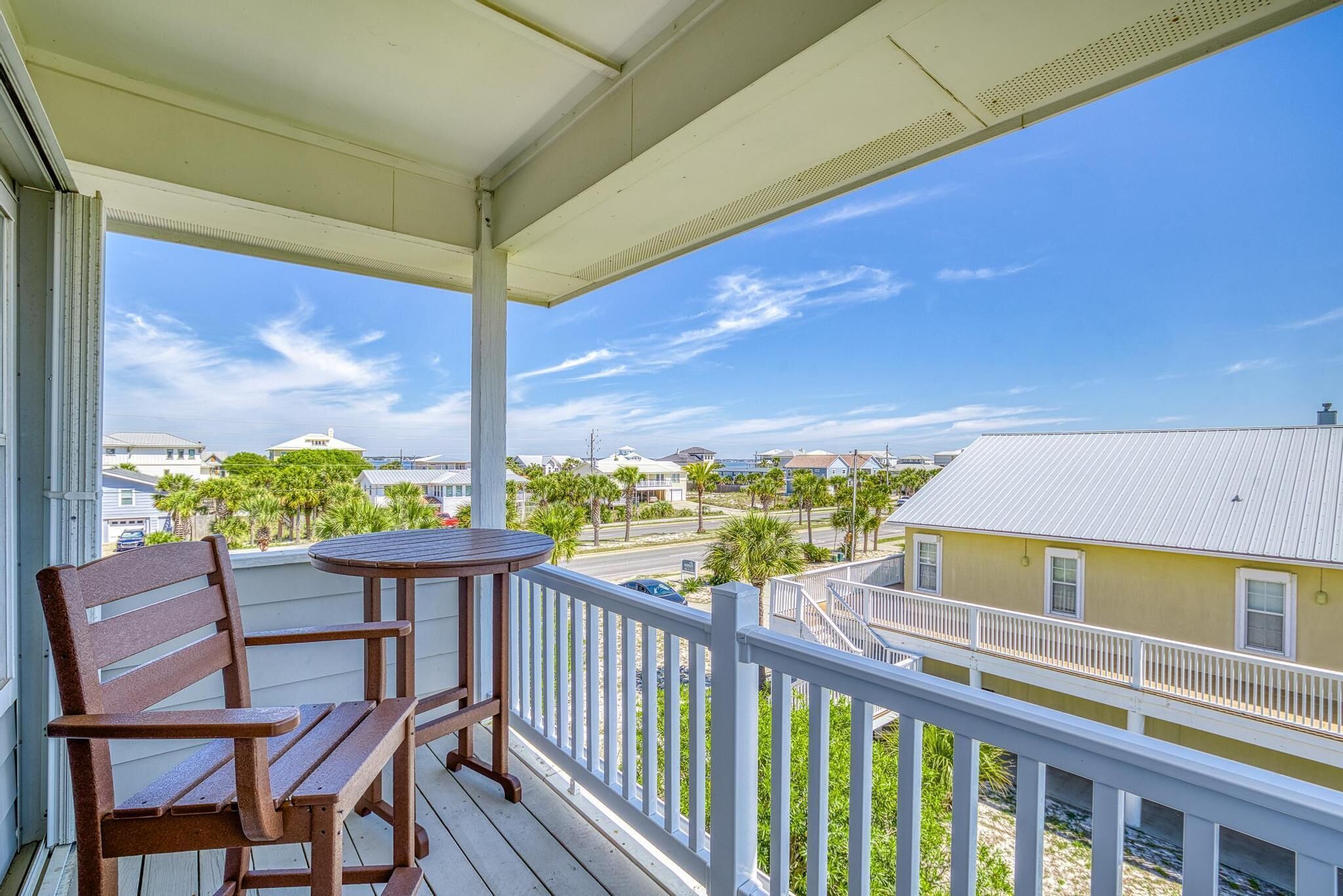 A balcony with a wooden table and chair, overlooking a residential area with houses and palm trees.