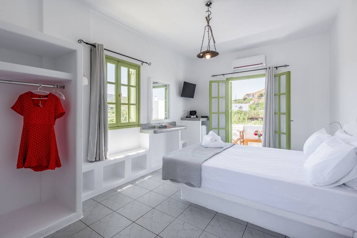 A hotel room with a bed, a TV, a window with a view, and a red dress hanging on a rack.
