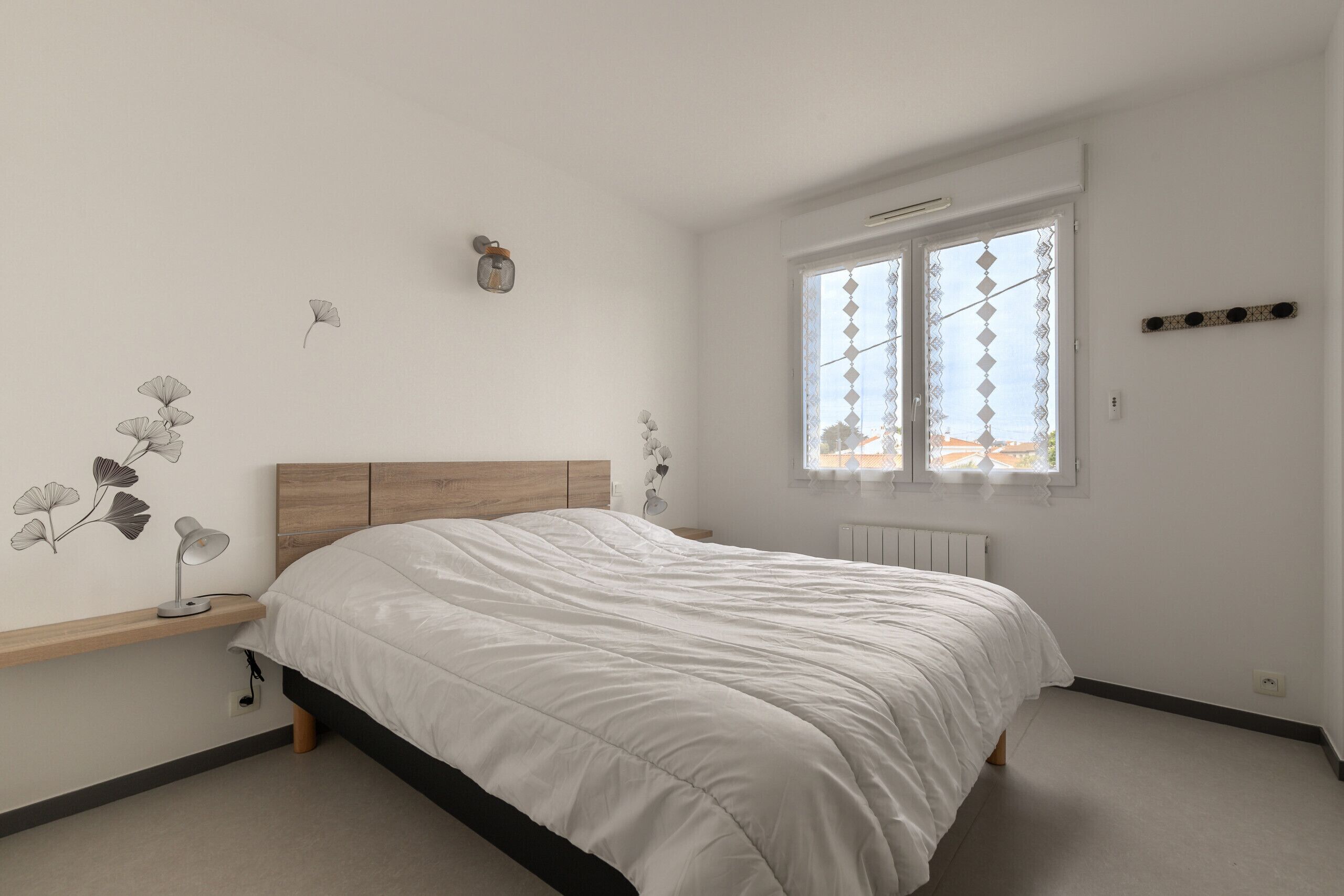 Een slaapkamer met een bed, een nachtkastje, een wandlamp en een raam met uitzicht op gebouwen.