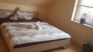Uma cama bem arrumada com colcha estampada e almofadas, uma mesa de cabeceira com abajur e uma janela com planta no parapeito.